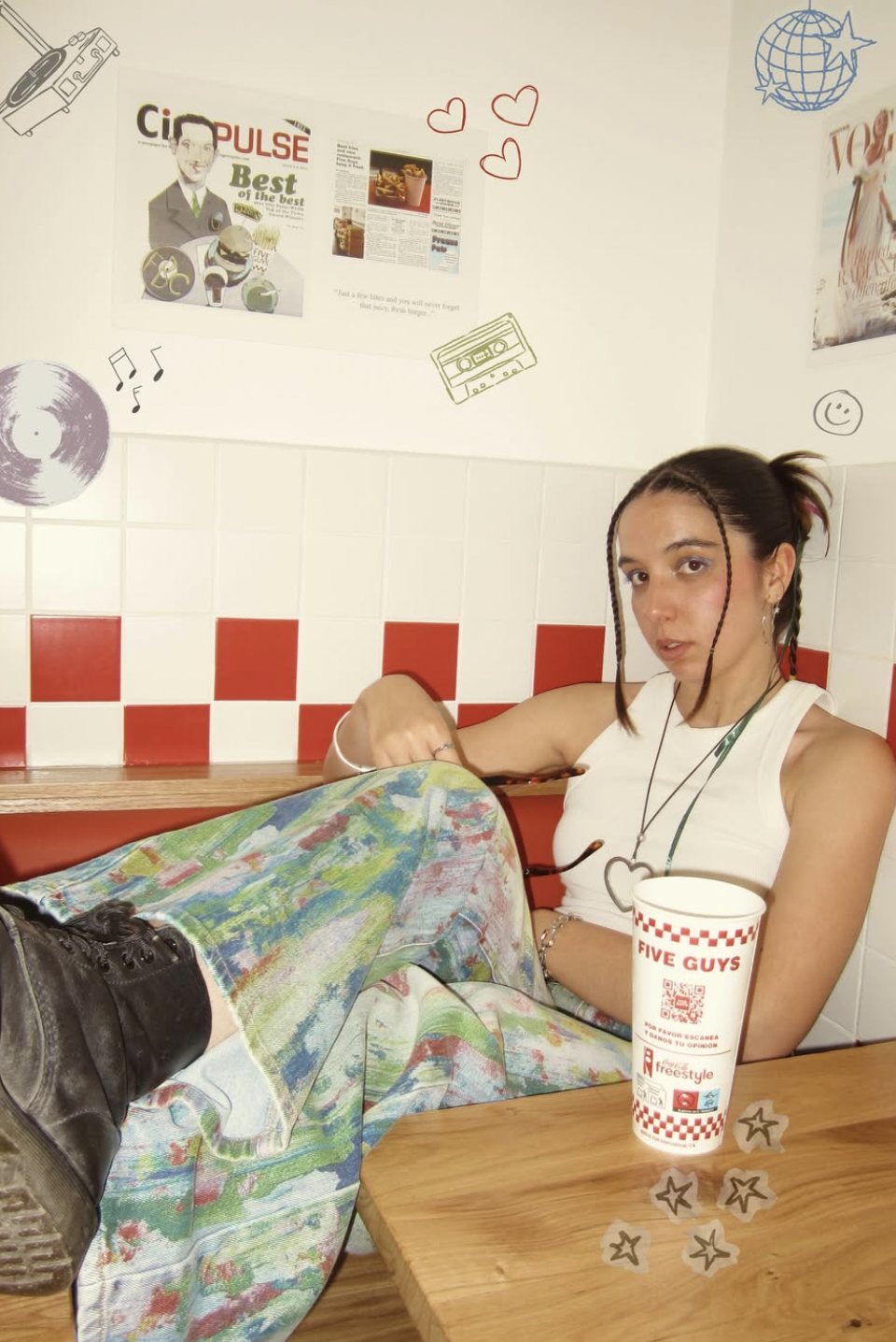 Juvika sitting in a diner booth, wearing a white top and painted trousers